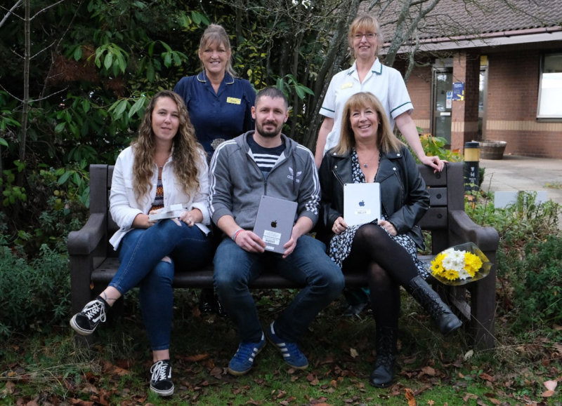 Kev Price’s family with ipads and sitting on the memorial bench. They are pictured with Di Evanson, Macmillan Rarer Tumour Clinical Nurse Specialist, and Emma Hicks, Macmillan Clinical Specialist Occupational Therapist